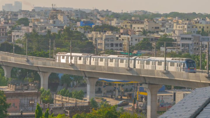 వీధిపై ఎత్తైన కాంక్రీట్ వంతెన మీద నడుస్తున్న తెల్లటి మూడు కోచ్‌ల మెట్రో రైలు, వెనుక నివాస భవనాలు.