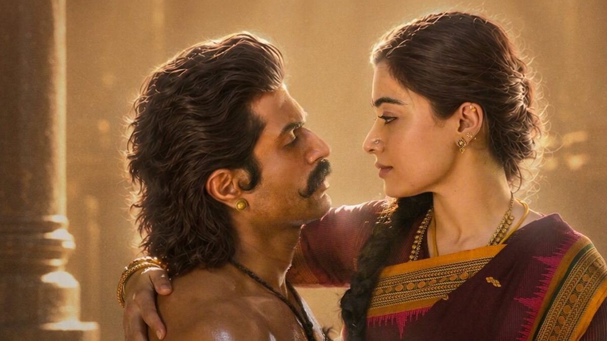 బంగారు వెలుతురులో రాతి స్తంభాల నేపథ్యంతో ఒకరినొకరు చూసుకుంటున్న విజయ్ దేవరకొండ, రష్మిక మందన్న; రష్మిక చేయి విజయ్ భుజంపై ఉంది.