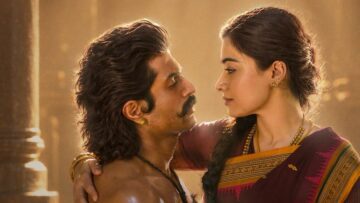 బంగారు వెలుతురులో రాతి స్తంభాల నేపథ్యంతో ఒకరినొకరు చూసుకుంటున్న విజయ్ దేవరకొండ, రష్మిక మందన్న; రష్మిక చేయి విజయ్ భుజంపై ఉంది.