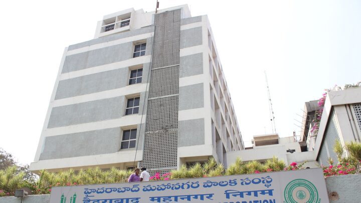 నీలి రంగు మూడు భాషల బోర్డు, ముందు పూల మొక్కలతో బహుళ అంతస్తుల గ్రేటర్ హైదరాబాద్ మున్సిపల్ కార్పొరేషన్ ప్రధాన కార్యాలయం.