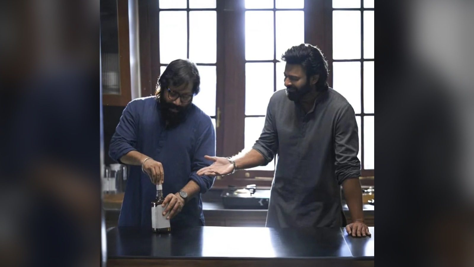 పొడవైన కిటికీలున్న వంటగదిలాంటి గదిలో ఇద్దరు వ్యక్తులు: నీలి చొక్కా, కళ్లద్దాలతో ఉన్న ఒకరు సాదా లేబుల్ ఉన్న సీసా మూత తీస్తుండగా, బూడిద రంగు కుర్తాలో ఉన్న మరొకరు నవ్వుతూ ఆయన వైపు చేయి చూపిస్తున్నారు.